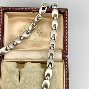 Puede incluir: Un brazalete plateado con una serie de eslabones ovalados y un cierre. Se muestra dentro de un joyero vintage con exterior granate e interior crema. El diseño del brazalete es sencillo y elegante, ideal para cualquier ocasión.