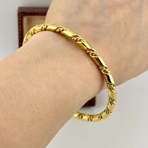 Op de afbeelding: Een goudkleurige armband met een gedraaid touwontwerp. De armband is gemaakt van een glanzend, reflecterend metaal en wordt om een pols gedragen. Het ontwerp heeft een reeks diagonale lijnen die een getextureerd uiterlijk creëren.
