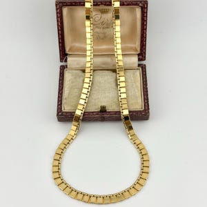 以下が含まれることがあります： 正方形のリンクデザインのゴールドカラーのネックレス。 ネックレスは、クリーム色の内装の開いたヴィンテージのマルーンのジュエリーボックスに表示されています。 ボックスには、ゴールドカラーの留め金とヒンジが付いています。