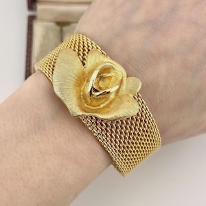 May include: A gold-tone cuff bracelet featuring a wide, flexible mesh band and a prominent, textured three-dimensional rose flower centrepiece. The metal surface has a brushed finish, creating a consistent golden appearance across the entire piece.