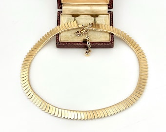 May include: A gold-toned metal necklace featuring a series of repeating, angled, chevron-like links arranged in a continuous circular band. The necklace includes a short adjustable chain extension with a small circular clasp at the end.