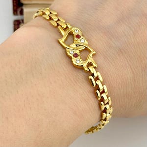 May include: Gold-coloured bracelet with a decorative centrepiece set with red and clear gemstones. The bracelet has a chain-link design. The centrepiece has a unique, stylized shape and is worn on a wrist.