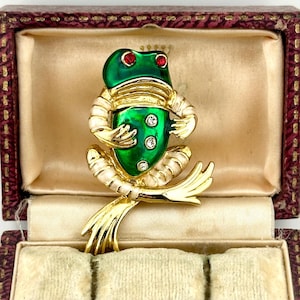 Puede incluir: Broche vintage de rana con cuerpo de esmalte verde, detalles dorados y ojos de pedrería roja. El broche presenta un diseño detallado con detalles de esmalte blanco y pedrería transparente. Se muestra en un joyero burdeos.