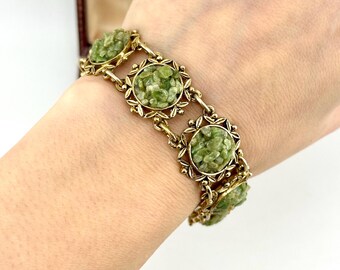 May include: A gold-tone link bracelet featuring circular settings, each containing a cluster of small, light green, textured stone chips. The bracelet is composed of multiple repeating segments connected by small metal links.
