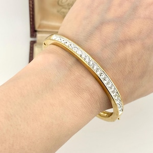 May include: A gold-toned hinged bangle bracelet featuring a single row of clear, round-cut crystals embedded along the centre of the band. The metal surface has a polished finish, and the design includes a secure clasp mechanism on the side.