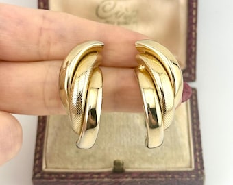 May include: A pair of gold-tone earrings featuring a layered, curved design. Each earring consists of three overlapping segments, alternating between a smooth, polished finish and a textured, cross-hatched pattern, creating a dimensional look.