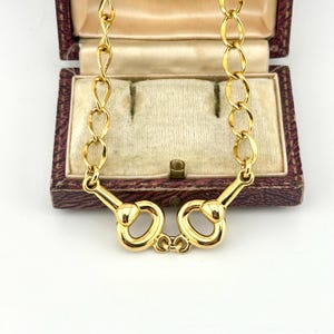 May include: A gold-toned chain necklace featuring a central horsebit-style metal link design. The necklace consists of polished oval-shaped links that connect to the horsebit centrepiece, which is composed of two symmetrical loops joined together.