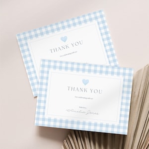Può includere: Due biglietti di ringraziamento a quadretti azzurri. Ogni biglietto ha un centro bianco con le parole "THANK YOU" e un cuore blu. I biglietti sono per festeggiare con qualcuno. Il nome "Amelia Jones" è scritto in basso.