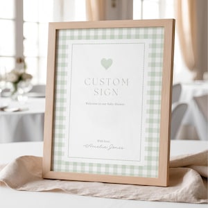 May include: A framed sign with a light wood frame. The sign has a white background with a pale green gingham border. The text reads "CUSTOM SIGN" and "Welcome to our Baby Shower" with a heart graphic.