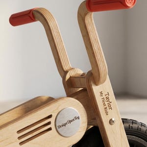 May include: A wooden balance bike with red handlebars. The bike features the text "Taylor My First Ride" and "Design Upon Toy". The bike has a silver emblem and black tires. The handlebars are approximately 10 inches wide.