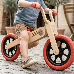 May include: A wooden balance bike with red tires and handlebars. The bike has a natural wood finish and black accents on the wheels. The child is wearing a blue shirt and brown shorts. The bike is designed for young children.