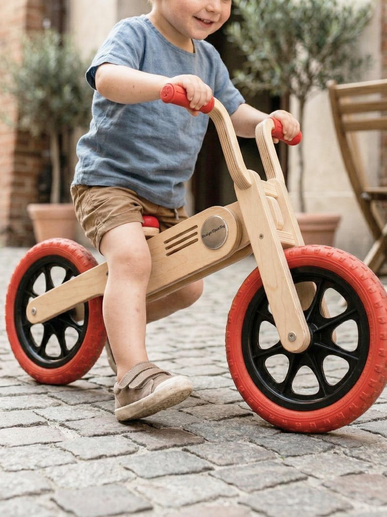 May include: A wooden balance bike with red tires and handlebars. The bike has a natural wood finish and black accents on the wheels. The child is wearing a blue shirt and brown shorts. The bike is designed for young children.