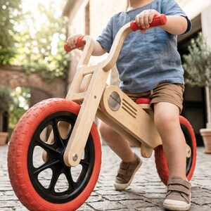 May include: A wooden balance bike with red tires and handlebars. The bike has a natural wood finish and black wheel spokes. The child-sized bike is designed for toddlers and young children. The bike is designed to help children learn balance and coordination.