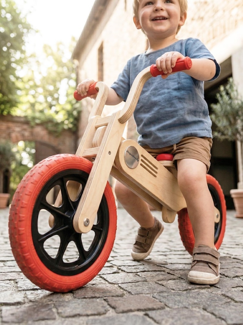 May include: A wooden balance bike with red tires and handlebars. The bike has a natural wood finish and black wheel spokes. The child-sized bike is designed for toddlers and young children. The bike is designed to help children learn balance and coordination.