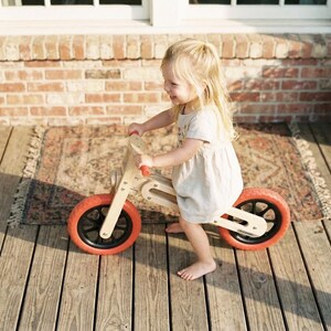 Op de afbeelding: Een houten loopfiets met oranje banden en zwarte velgen. De fiets heeft een natuurlijke houten afwerking en rode handvatten. Een kind in een lichte jurk rijdt op de fiets op een houten terras.