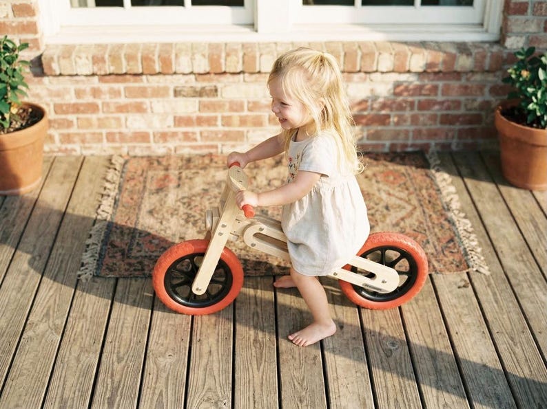 Op de afbeelding: Een houten loopfiets met oranje banden en zwarte velgen. De fiets heeft een natuurlijke houten afwerking en rode handvatten. Een kind in een lichte jurk rijdt op de fiets op een houten terras.
