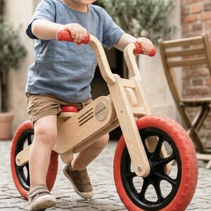 May include: A wooden balance bike with red tires and handlebars. The bike has a natural wood finish and a small red seat. The wheels have black spokes. The child is wearing a blue shirt and brown shorts.