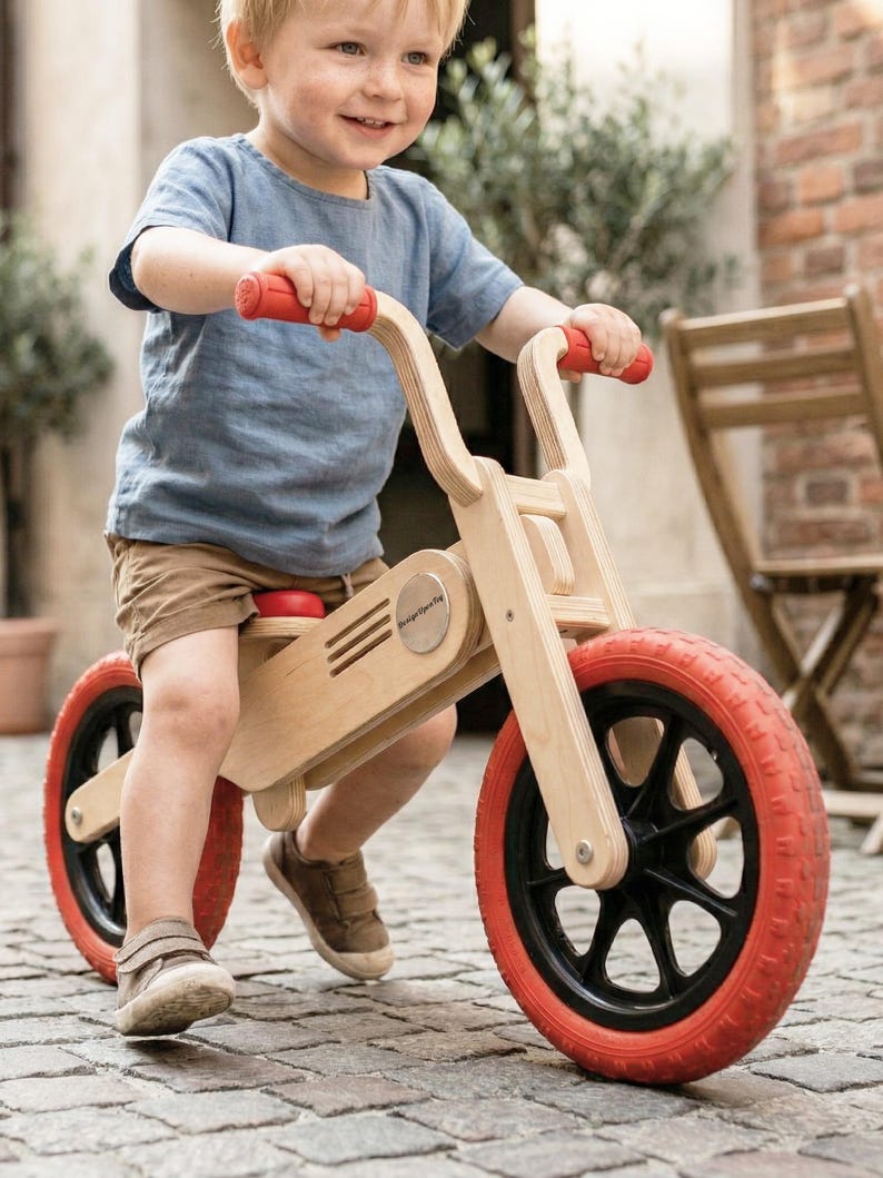 May include: A wooden balance bike with red tires and handlebars. The bike has a natural wood finish and a small red seat. The wheels have black spokes. The child is wearing a blue shirt and brown shorts.