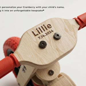 May include: Close-up of a wooden scooter with red handlebars and wheels. The scooter's wooden surface is engraved with the name "Lillie" and the date "7.14.2024".