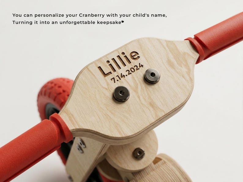 May include: Close-up of a wooden scooter with red handlebars and wheels. The scooter's wooden surface is engraved with the name "Lillie" and the date "7.14.2024".