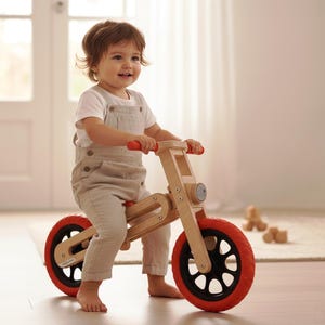 May include: A wooden balance bike with red tires and handlebars. The bike has a natural wood finish and black accents. The child is wearing a white shirt and tan overalls. The bike is designed for toddlers.