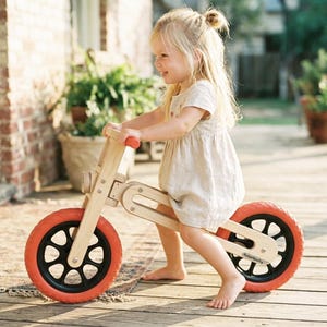 Op de afbeelding: Een houten loopfiets met rode banden en zwarte spaken. De fiets heeft een natuurlijke houten afwerking en is ontworpen voor jonge kinderen. Het kind draagt een lichtgekleurde jurk en lacht terwijl het op de fiets rijdt.