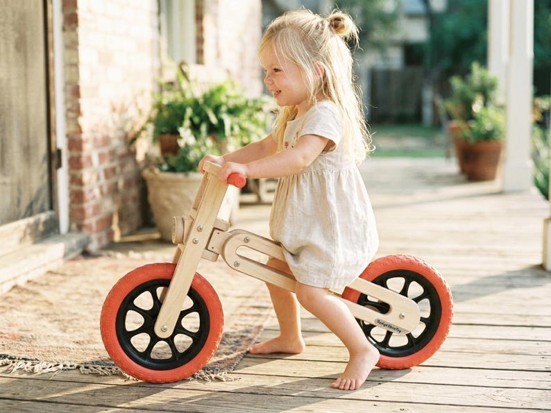 Op de afbeelding: Een houten loopfiets met rode banden en zwarte spaken. De fiets heeft een natuurlijke houten afwerking en is ontworpen voor jonge kinderen. Het kind draagt een lichtgekleurde jurk en lacht terwijl het op de fiets rijdt.