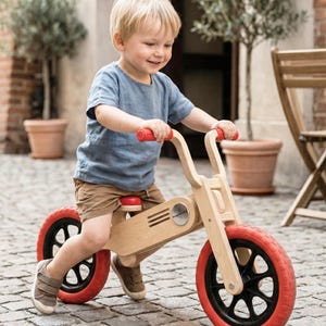 May include: A wooden balance bike with red tires and handlebars. The bike has a natural wood finish and black wheels. The child is wearing a blue shirt and brown shorts. The bike is designed for toddlers and young children.