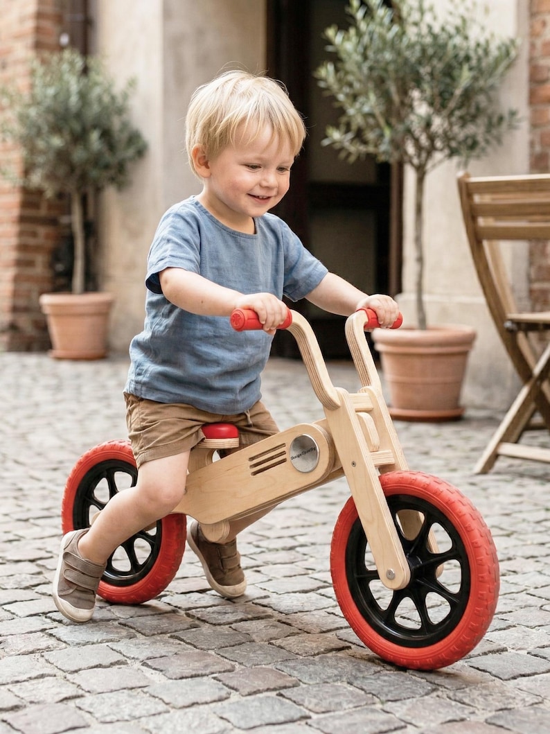 May include: A wooden balance bike with red tires and handlebars. The bike has a natural wood finish and black wheels. The child is wearing a blue shirt and brown shorts. The bike is designed for toddlers and young children.