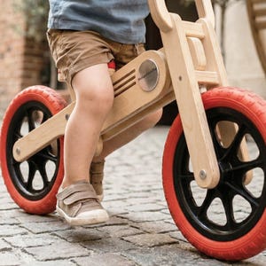 May include: A wooden balance bike with red tires and black rims. The bike has a natural wood finish and red handlebars. The child is wearing brown shorts and a blue shirt. The bike is on a cobblestone street.