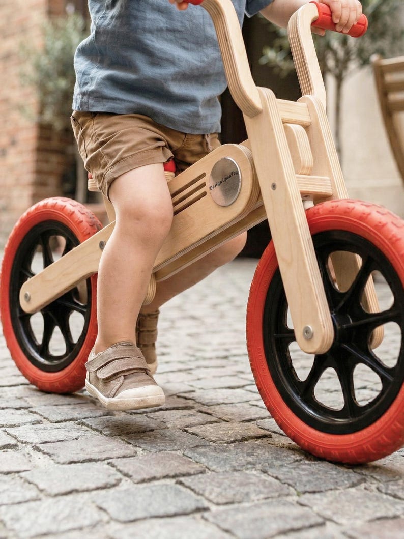 May include: A wooden balance bike with red tires and black rims. The bike has a natural wood finish and red handlebars. The child is wearing brown shorts and a blue shirt. The bike is on a cobblestone street.