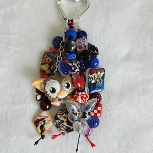 May include: A keychain featuring a heart-shaped silver ring attached to a cluster of colorful beads and charms. The charms include a small figurine of a creature with large ears, a rectangular Gremlins movie tag, and a butterfly-shaped bead.