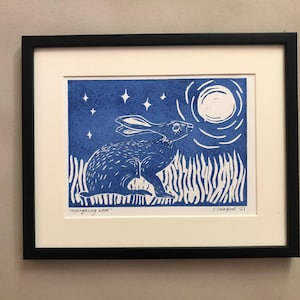 May include: Framed linocut print in blue and white, depicting a hare under a full moon. The artwork includes stars, tall grass, and the text "moongazing hare". The print is set in a black frame.