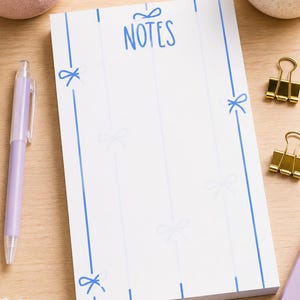 Può includere: Un blocco note rettangolare bianco con un bordo blu decorato da illustrazioni di fiocchi ripetuti e la parola NOTES stampata in blu in alto. Il blocco note presenta linee verticali blu e motivi a fiocco azzurri nella parte centrale.