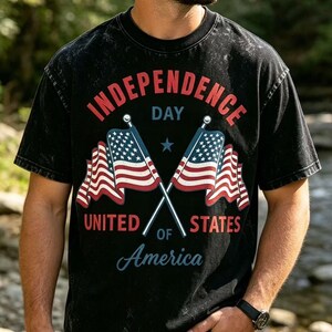 Puede incluir: Una camiseta negra con efecto lavado al ácido que presenta un gráfico de dos banderas estadounidenses cruzadas. El texto dice INDEPENDENCE DAY UNITED STATES OF AMERICA en letras rojas y azules. Incluye una pequeña estrella azul sobre las banderas.