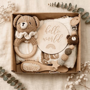 May include: A baby gift set in a cardboard box. The set includes a crochet bear rattle, a wooden sign with "hello world" text, a pacifier clip with a crochet bear, a wooden brush, and a muslin blanket. The items are in neutral colours.