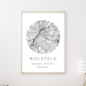 Könnte beinhalten: Ein Schwarzweißdruck einer Stadtkarte mit dem Städtenamen "BIELEFELD" und den Koordinaten "53° 37' 12" N 27° 19' 44"E" und dem Wort "GERMANY" darunter. Der Druck ist gerahmt und hängt an einer weißen Wand über einem Holztisch.