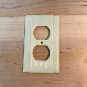 Könnte beinhalten: Eine hellcremefarbene Steckdosenabdeckung mit zwei ovalen Öffnungen und einem kleinen kreisförmigen Loch. Die Abdeckung hat ein vertikal gestreiftes Muster in der Mitte und einen glatten, rechteckigen Rand. Der Hintergrund ist eine hellfarbige Holzoberfläche.