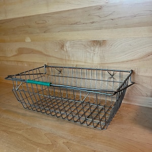 May include: A silver wire dish rack with a green plastic handle. The rack is sitting on a wooden surface.