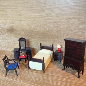 Puede incluir: Juego de muebles de dormitorio en miniatura en color marrón oscuro. Incluye una cama, una cómoda, un tocador con espejo, una mesita de noche con lámpara y una silla. La cama tiene una colcha blanca.