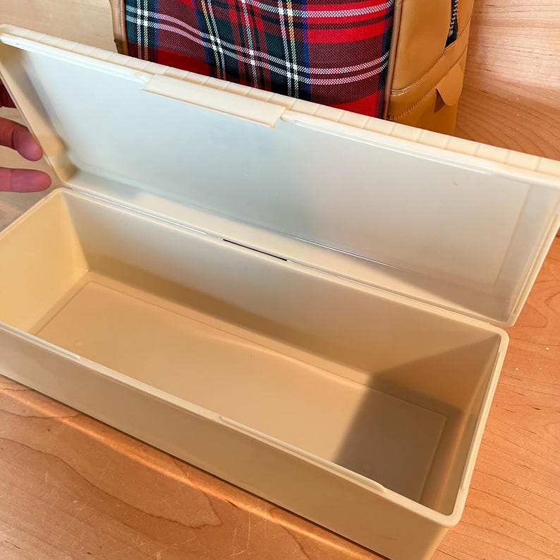 May include: An open, rectangular, cream-coloured plastic storage box with a hinged lid. The box is empty and sits on a wooden surface. A plaid pattern bag is visible in the background.