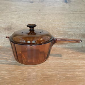 May include: A translucent amber-coloured glass saucepan with a matching lid and a long handle. The pot features a vertical ribbed texture along the exterior walls and a circular knob on the centre of the lid, constructed from heat-resistant glass.