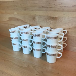 Puede incluir: Una pila de tazas de cerámica blanca con diseños florales marrones. Las tazas están apiladas en un patrón escalonado, con las asas alternando lados.