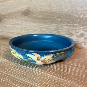 Puede incluir: Un cuenco redondo de cerámica azul con un diseño floral. El cuenco tiene un borde elevado y está decorado con flores amarillas y blancas similares a lirios. El cuenco probablemente se utiliza con fines decorativos o para servir objetos pequeños.