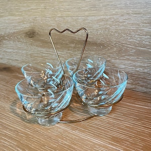 May include: A set of five clear glass bowls with a decorative swirl pattern, held in a gold-toned metal carrier with a heart-shaped handle. The bowls are designed for serving snacks or appetizers.