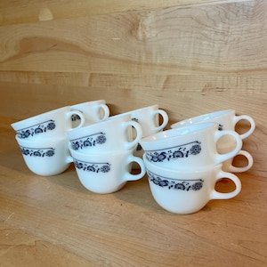 Puede incluir: Un juego de ocho tazas de cerámica blanca con un patrón floral azul. Las tazas están apiladas en dos filas de cuatro.