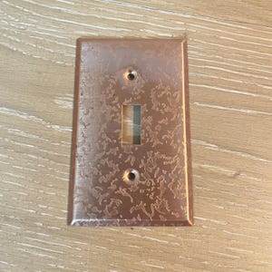 May include: A copper-colored light switch cover with a textured surface. The cover has a rectangular shape with a single switch opening and two screw holes. The copper finish has a mottled appearance.