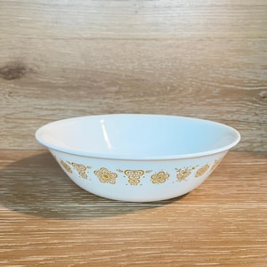 Puede incluir: Cuenco blanco con un patrón floral dorado alrededor del borde. El cuenco es redondo y tiene un diseño simple y elegante. El fondo es una superficie de madera marrón claro.