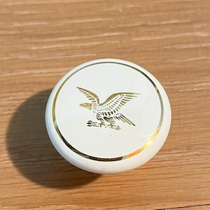 May include: A white, round cabinet knob with a gold eagle design. The eagle is centered on the knob and is surrounded by a gold border. The knob is likely made of ceramic or porcelain.