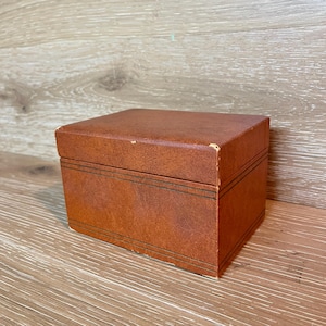 May include: A rectangular brown box with a textured surface and a lid. The exterior features thin, dark horizontal lines etched around the perimeter of the base and the lid, with visible wear on the top corner edges of the box.